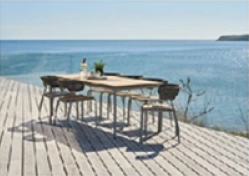 Angled lifestyle shot of the Mindo 101 outdoor chair showing teak seat and woven rope backrest, a large matching table, with an ocean backdrop on a sleek infinity deck.