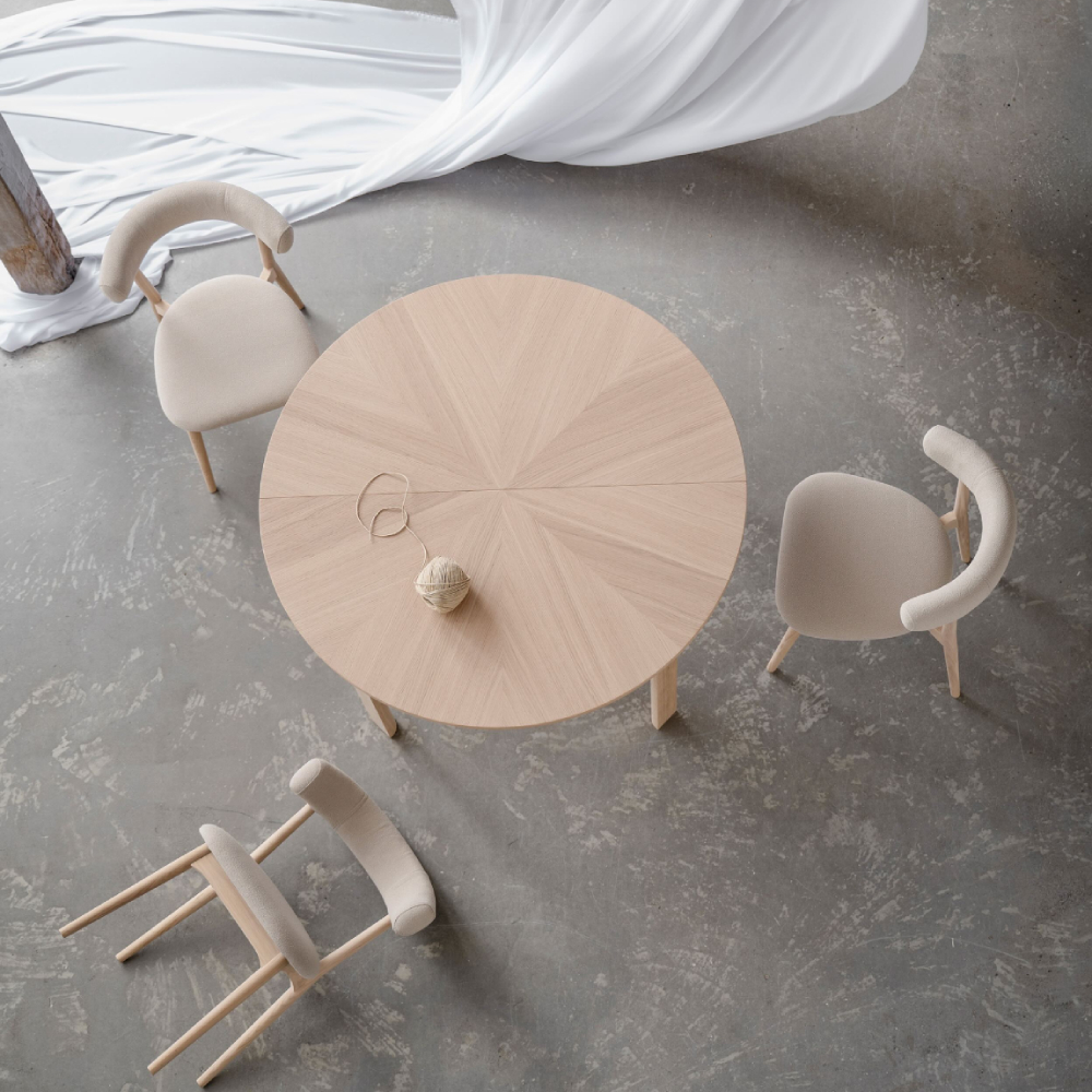 Top-down view of a round light oak dining table with four upholstered Aro dining chairs arranged on a polished concrete floor in a bright studio space.