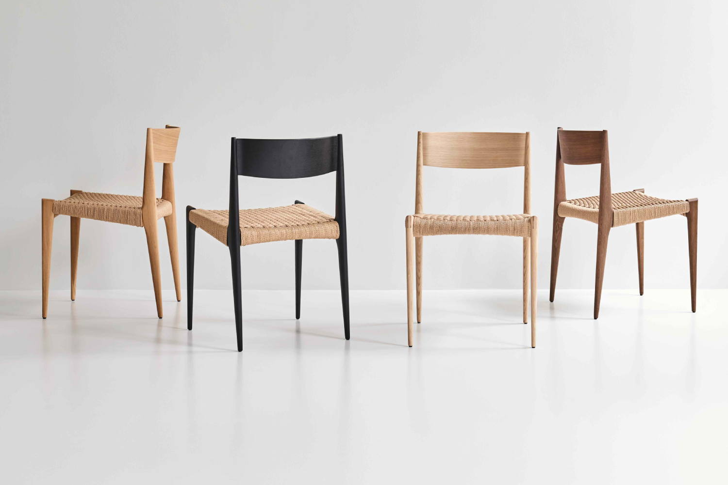 Four DK3 Pia Chairs in assorted oak finishes with woven seats, arranged in a minimal white studio setting.