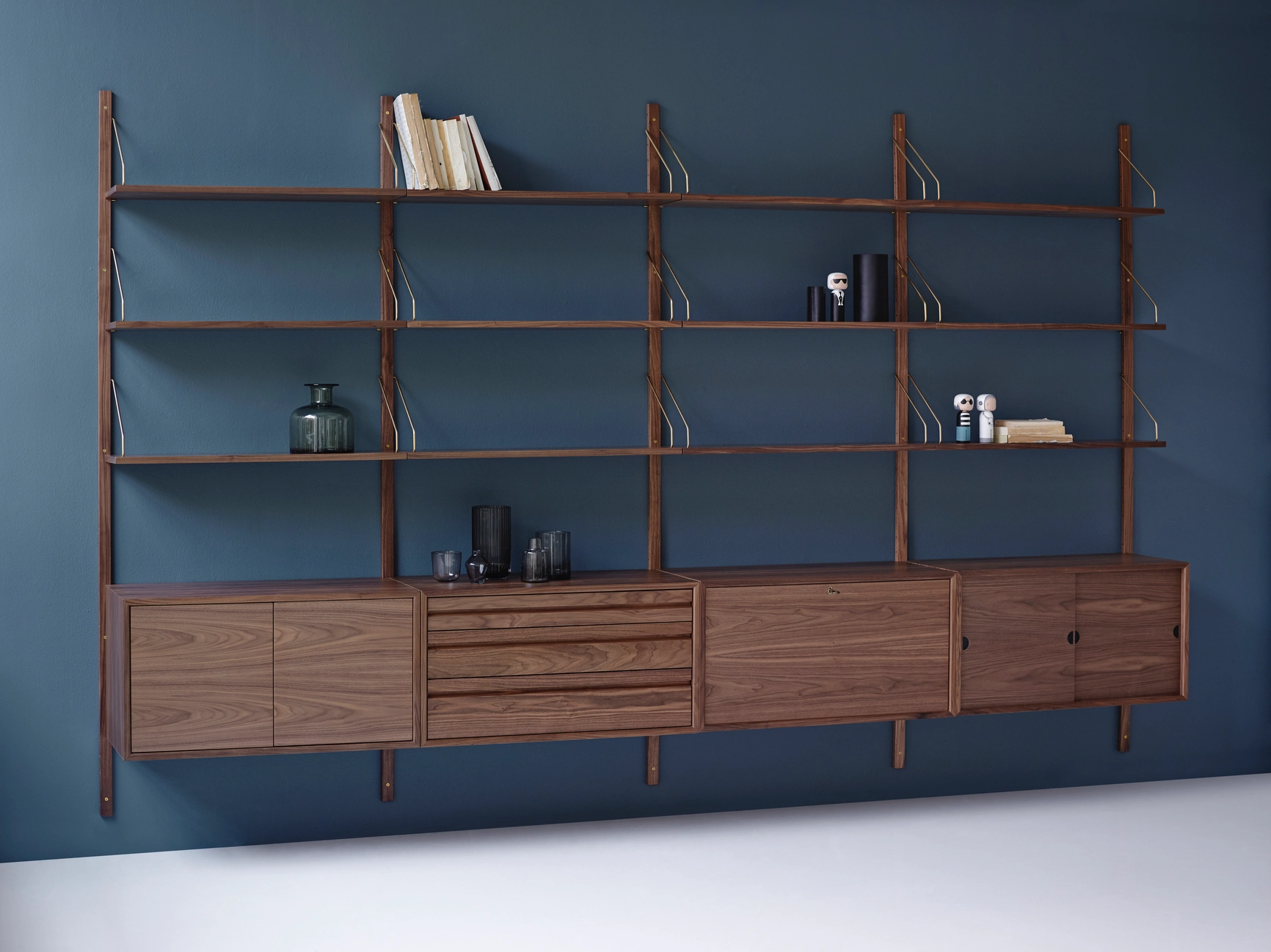 DK3 Royal System shelving in walnut with cabinets and open shelves, mounted on a blue wall and styled with books and glassware.