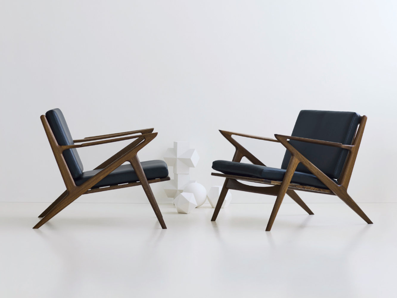 Two DK3 Z-Chairs in smoked oak with black leather upholstery, shown facing each other in a minimal white studio setting.